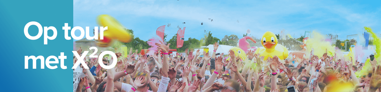 Een uitbundige festivalmenigte onder een blauwe lucht, gooit kleurpoeder in de lucht met een grote opblaasbare badeend in het midden, tijdens de zomertour van X²O badkamers.